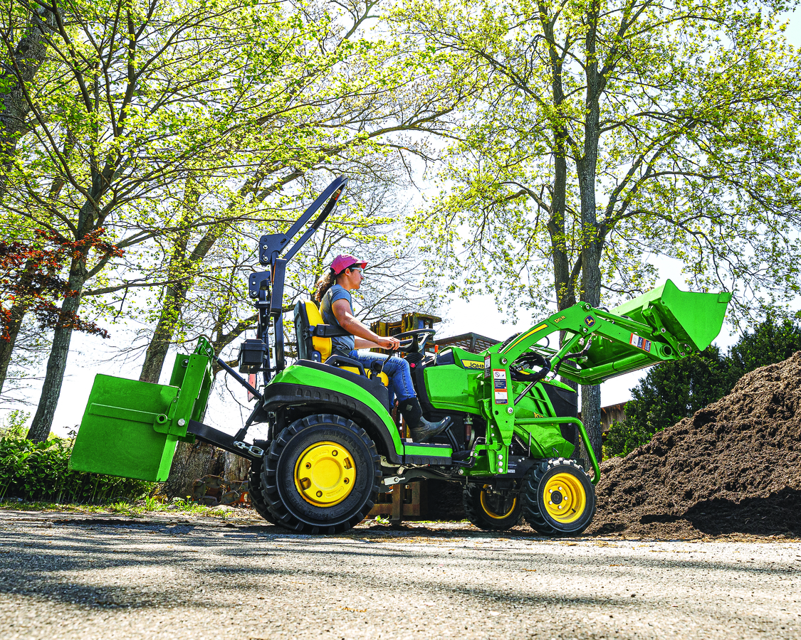A young woman operates a bright green John Deere 1 Series tractor, equipped with a front loader and 