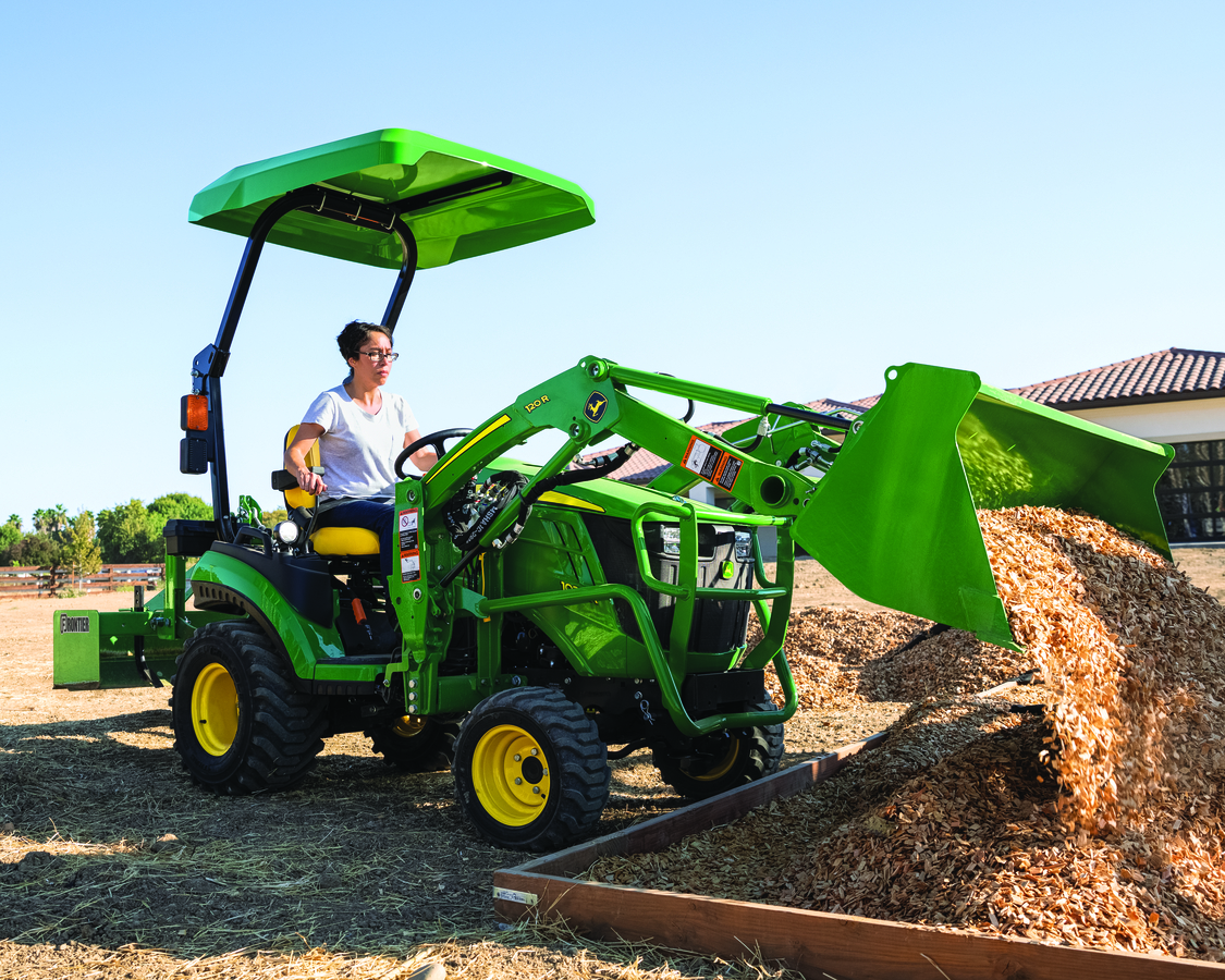 A woman in glasses and a white t-shirt operates a green John Deere compact utility tractor with a fr