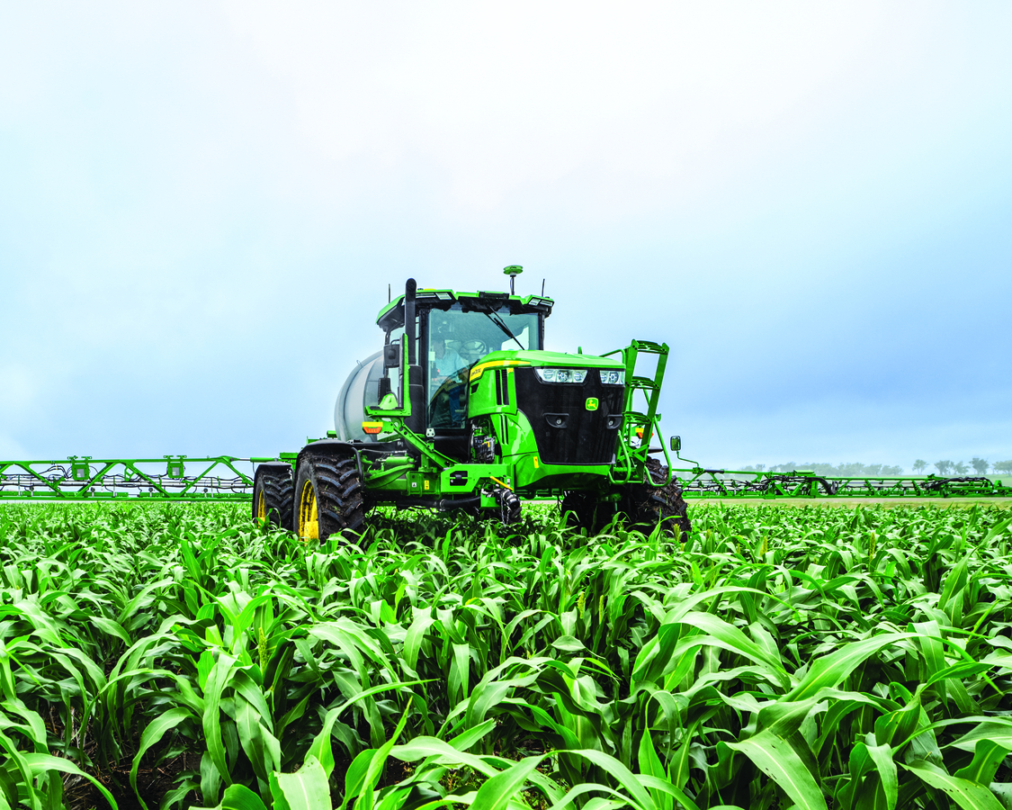A large, green John Deere self-propelled sprayer with a prominent front-mounted boom is shown from a