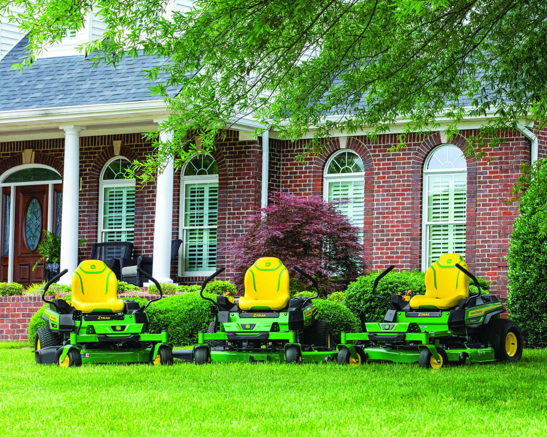 Three green and yellow John Deere zero-turn lawn mowers are parked side-by-side on a vibrant green l